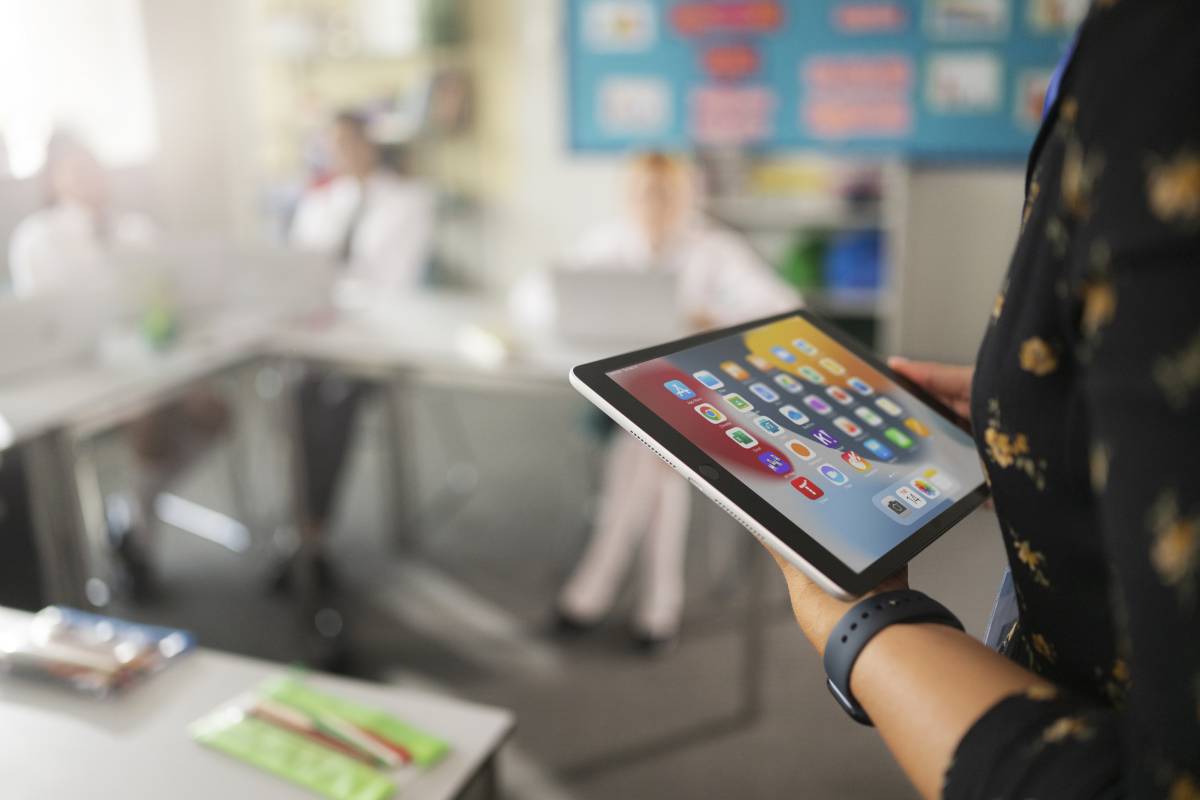 Ein iPad mit App Icons am Display, gehalten von einer Lehrerin in einem Klassenzimmer, im Hintergrund Schüler:innen