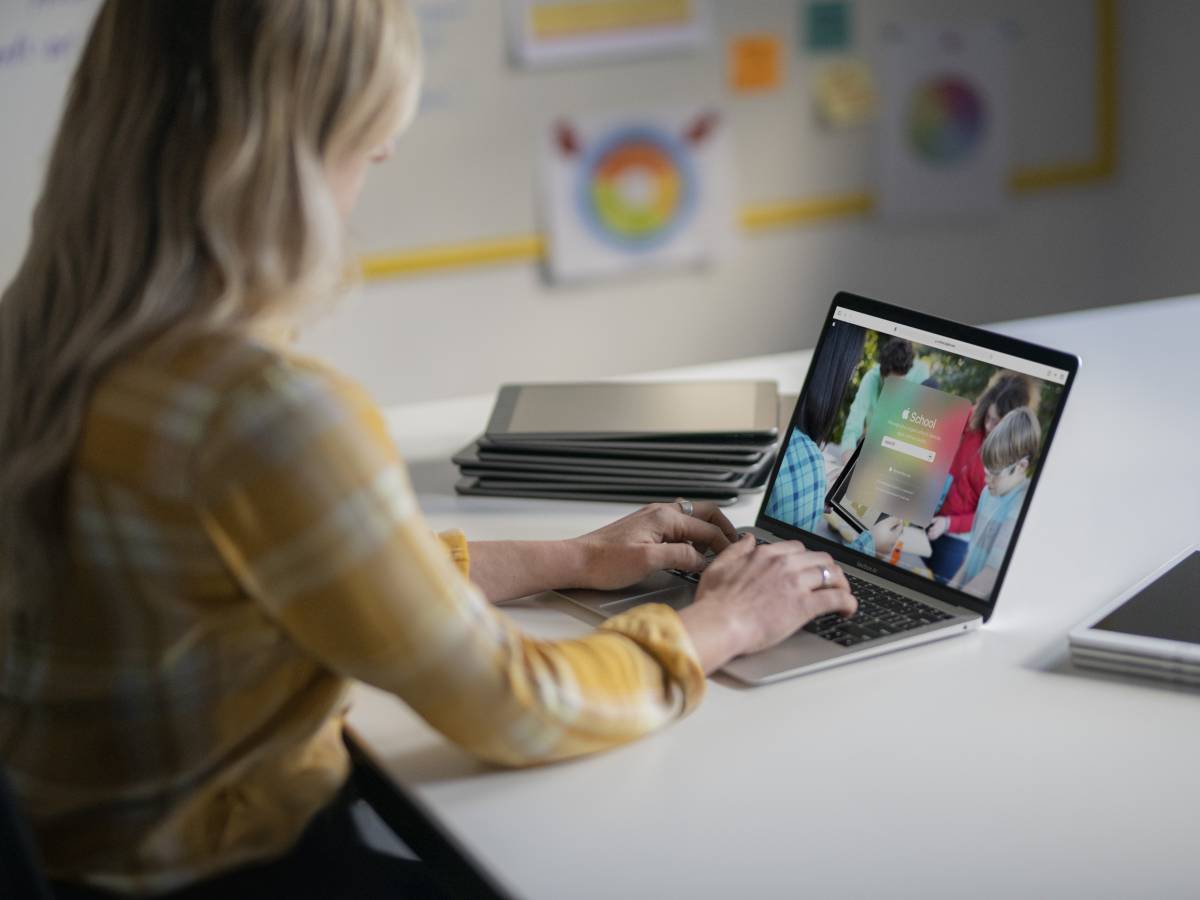 : Eine IT-Administratorin verwaltet auf einem MacBook die Geräte ihrer Schule, im Hintergrund ein Stapel iPads