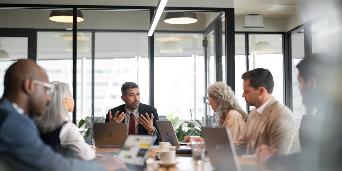Sechs Personen bei einem Meeting in einer Bildungseinrichtung, am Tisch stehen mehrere MacBooks
