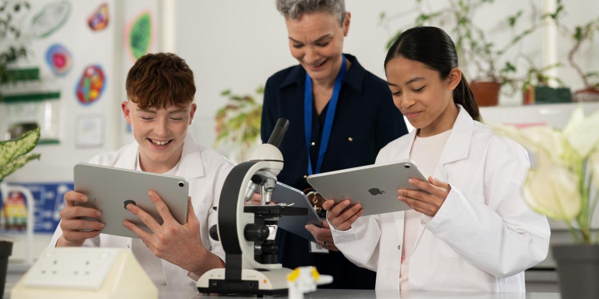 Eine Lehrerin steht mit einer Schülerin und einem Schüler in einem Chemielabor, alle drei arbeiten am iPad